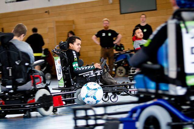 Mehrere Jugendliche in elektrischen Rollst&uuml;hlen spielen Fu&szlig;ball in einer Sporthalle, ein Ball rollt zwischen ihnen.