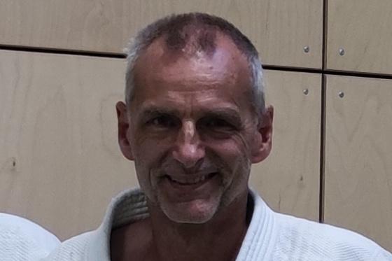 Lächelnder Mann in einem weißen Judogi vor einer Holzwand, Teil einer Gruppe von Judokas.