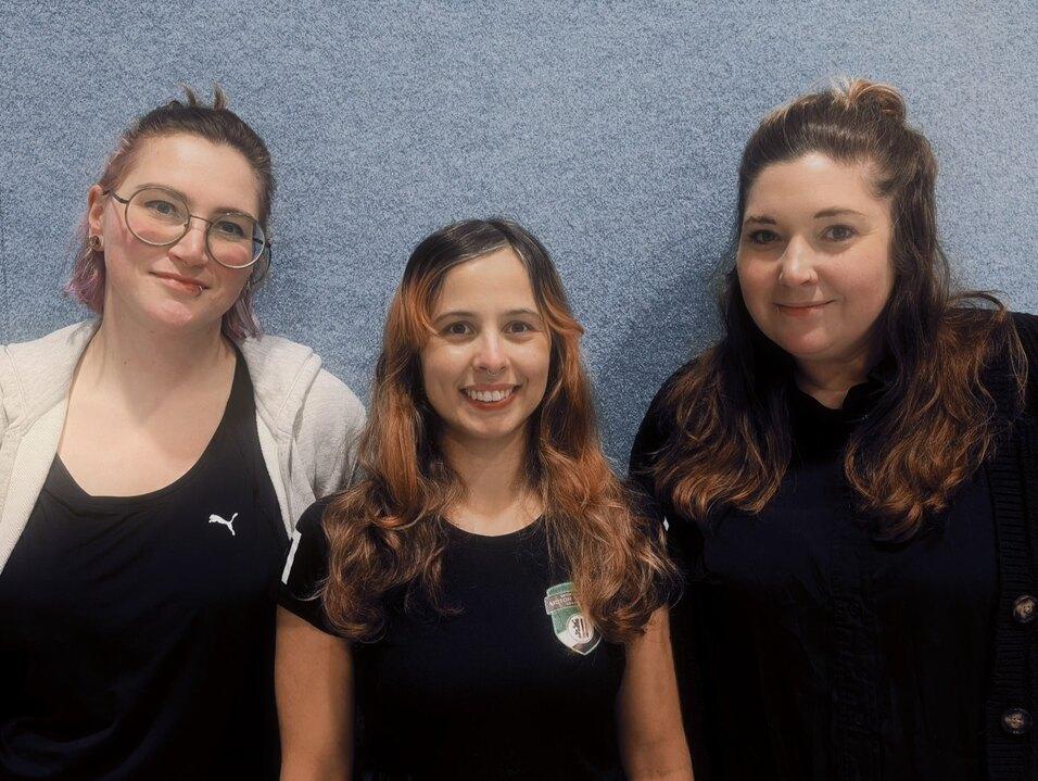 Drei Frauen stehen vor einer blauen Wand, lächeln und tragen schwarze Kleidung mit verschiedenen Frisuren.