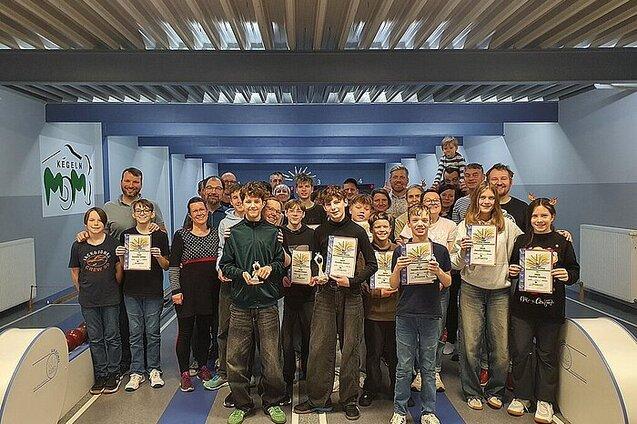 Gruppe von Kindern und Erwachsenen mit Pokalen und Urkunden in einer Kegelbahn, fr&ouml;hliche Stimmung im Hintergrund.