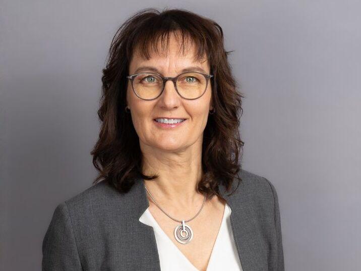 Porträt einer lächelnden Frau mit braunen Haaren, Brille und grauem Blazer vor einem grauen Hintergrund.