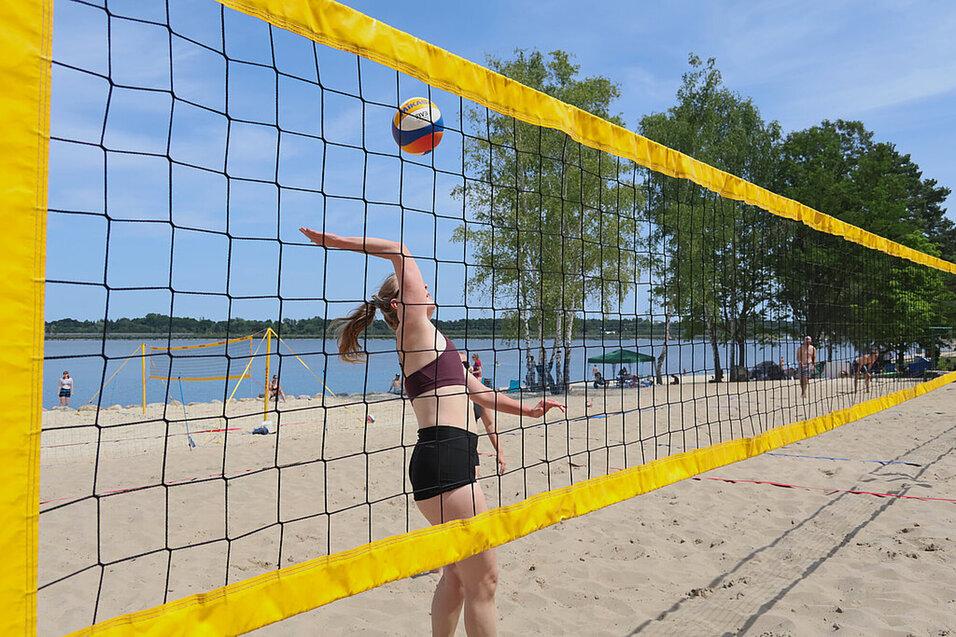 Spielerin im Sand wirft einen Volleyball &uuml;ber das Netz an einem Strand mit Wasser im Hintergrund.