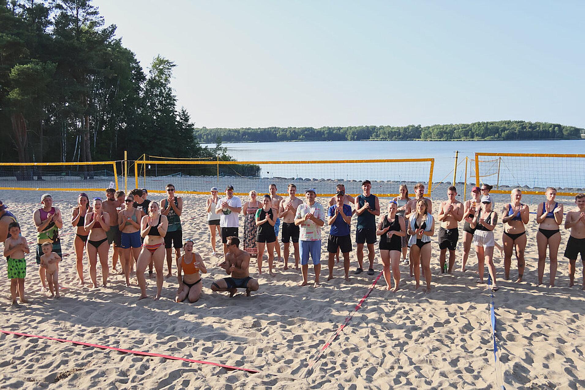 Gruppe von Menschen in Strandsportbekleidung steht am Sandstrand vor einem See und Volleyballnetzen.