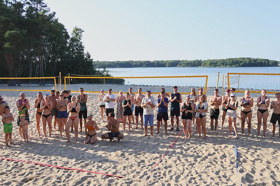Gruppe von Menschen in Strandsportbekleidung steht am Sandstrand vor einem See und Volleyballnetzen.
