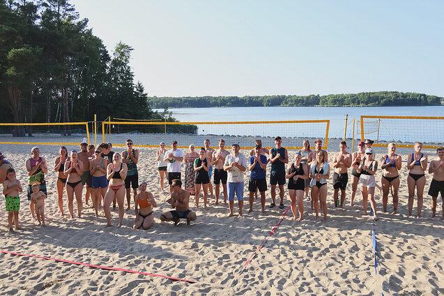 Gruppe von Menschen in Strandsportbekleidung steht am Sandstrand vor einem See und Volleyballnetzen.