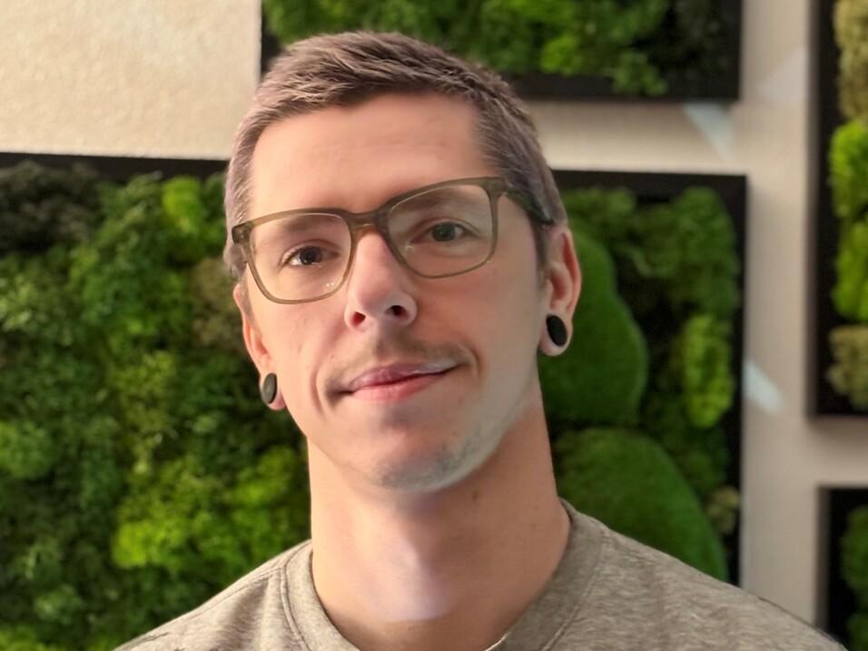 Porträt eines Mannes mit Brille und lässigem Outfit vor einer Wand mit grünen Pflanzenarrangements.