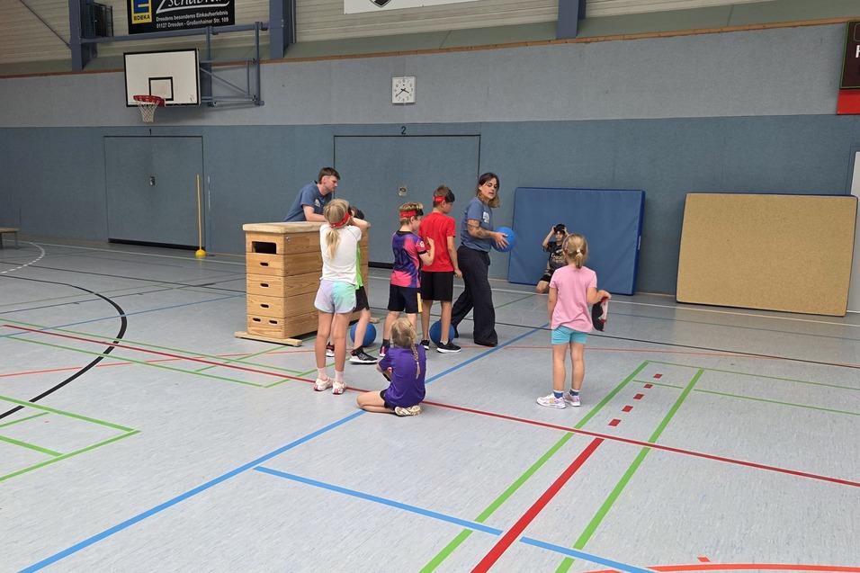 Gruppe von Kindern beim Spielen in einer Sporthalle mit Kasten, B&auml;llen und farbigen Linien auf dem Boden.