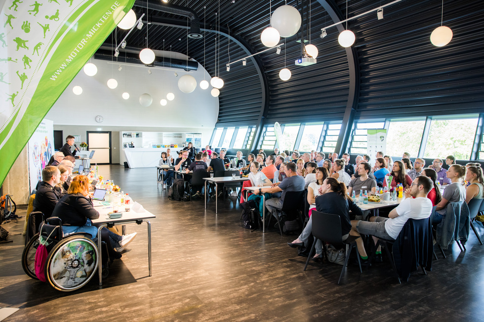 Konferenz mit Publikum in modernem Raum; Panel mit Rednern vorne, Tische mit Getr&auml;nken und Snacks.