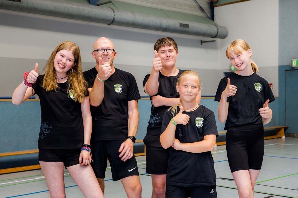 F&uuml;nf Personen in sportlichen Schwarz-Shirts zeigen Daumen hoch in einer Sporthalle.