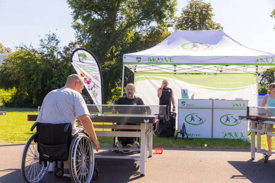 Zwei Tischtennisspieler im Rollstuhl spielen an Tischen vor einem Informationsstand im Freien.