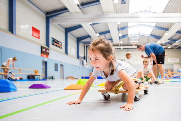Junge, die auf einem Rollbrett in einer Sporthalle kriecht, umgeben von bunten Trainingsger&auml;ten und anderen Kindern.