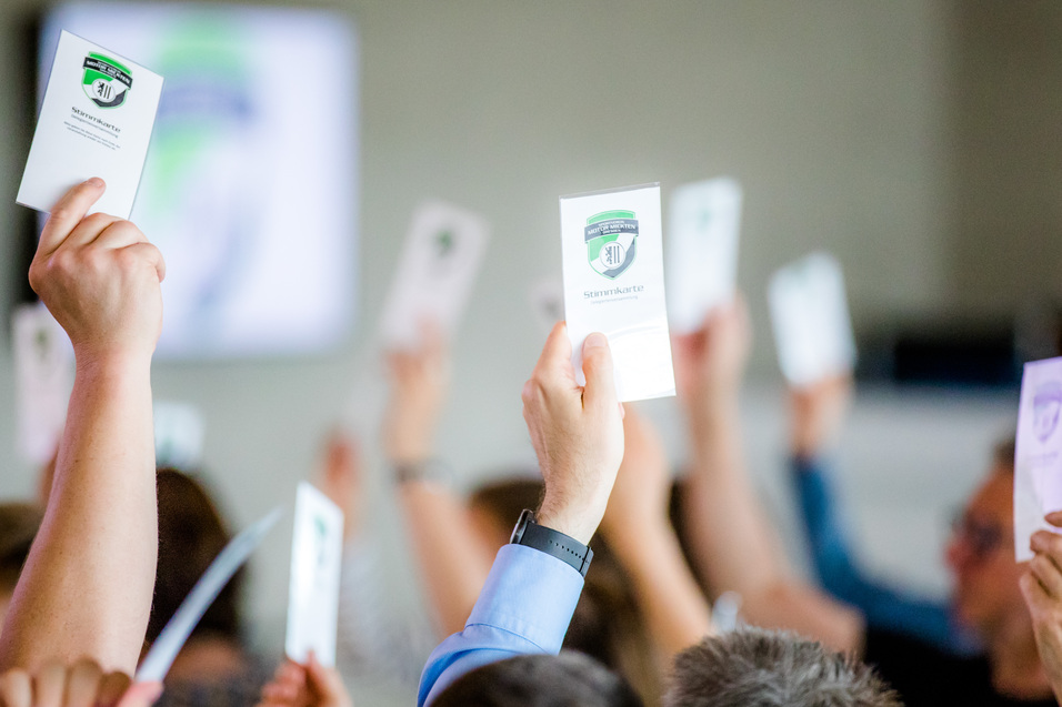 H&auml;nde mit Stimmkarten heben sich in einem Versammlungssaal, im Hintergrund ein verschwommenes Bildschirmbild.