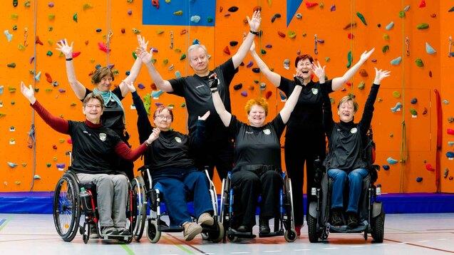 Gruppe von Menschen im Rollstuhl, die vor einer bunten Kletterwand fr&ouml;hlich posieren und die H&auml;nde heben.