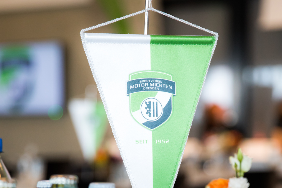 Tisch mit einem Wimpel des Sportvereins Motor Mickten, Getr&auml;nken und einer Blumenvasen im Hintergrund.
