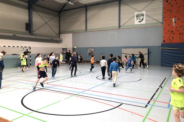 Gruppe von Kindern und Jugendlichen in Sportkleidung spielt in einer gro&szlig;en Turnhalle mit Basketballkorb und Kletterwand.