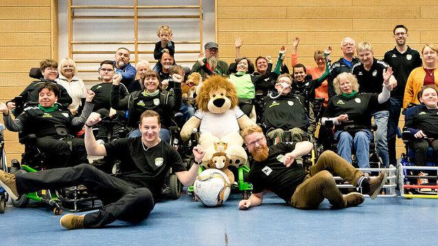 Gruppe von Menschen in Rollst&uuml;hlen und Stehenden, die gemeinsam lachen und jubeln, umgeben von einem gro&szlig;en Pl&uuml;schl&ouml;wen.