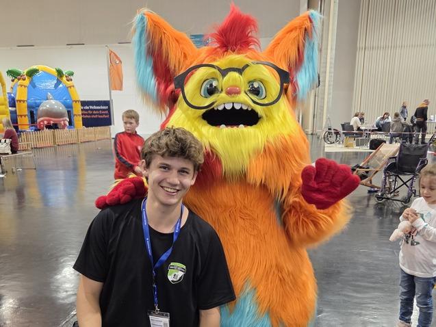 Ein junger Mann kniet neben einem bunten, felligem Maskottchen mit Brille auf einer Veranstaltungshalle.