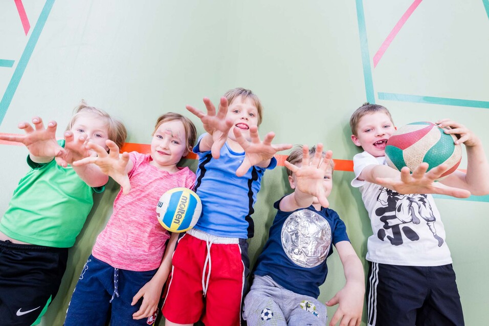 F&uuml;nf Kinder liegen auf dem Boden einer Sporthalle und strecken H&auml;nde zu Volleyball und Basketball.