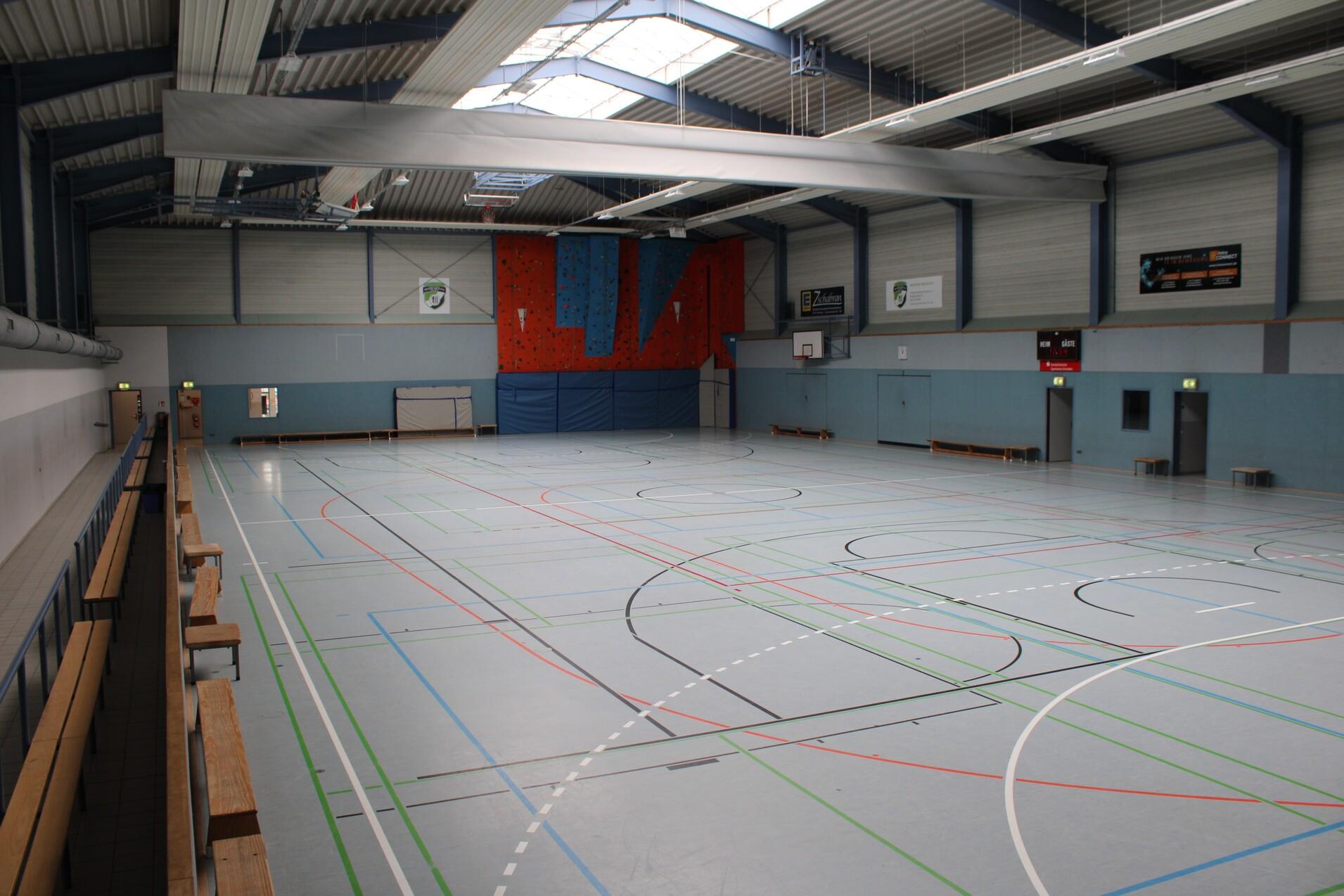 Gro&szlig;e Sporthalle mit gr&uuml;ner Bodenmarkierung, Trib&uuml;ne und roten W&auml;nden im Hintergrund. Tageslicht durch Fenster.