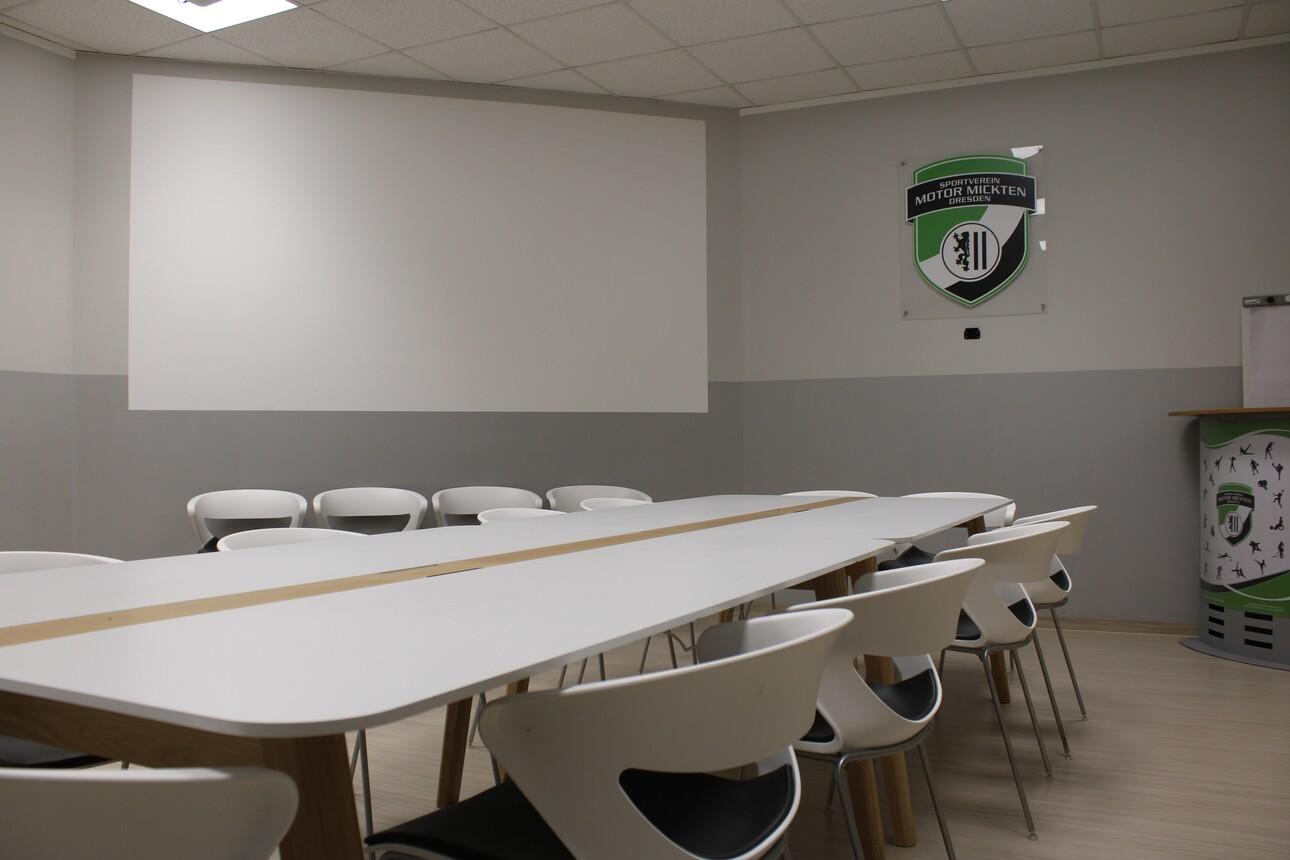 Konferenzraum mit einem langen Tisch und mehreren St&uuml;hlen, grauen W&auml;nden und einem Wandlogo auf der rechten Seite.