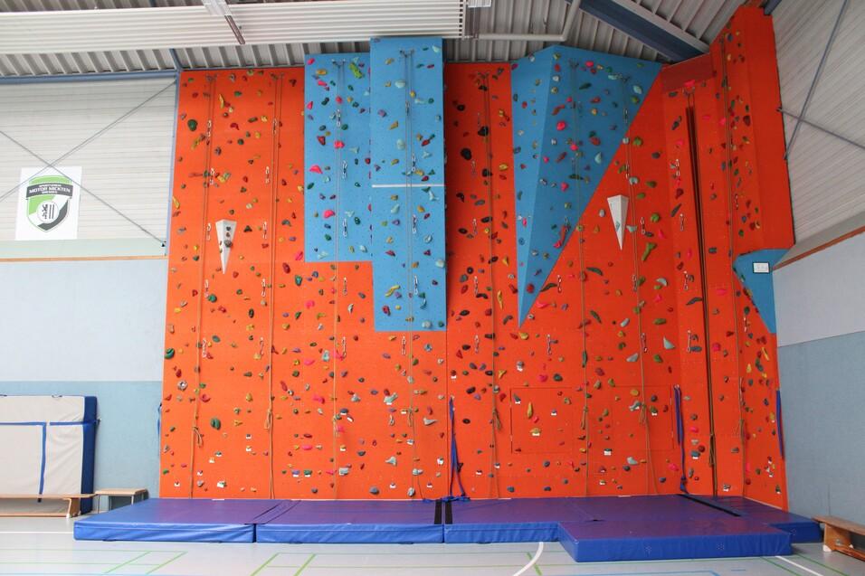 Bunte Kletterwand in einer Turnhalle mit verschiedenen Griffen und Matten im Vordergrund.