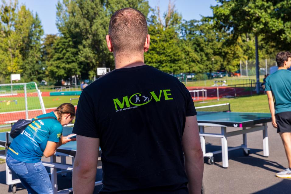 R&uuml;ckansicht einer Person in einem schwarzen T-Shirt mit der Aufschrift "MOVE", umgeben von Freizeitaktivit&auml;ten im Freien.