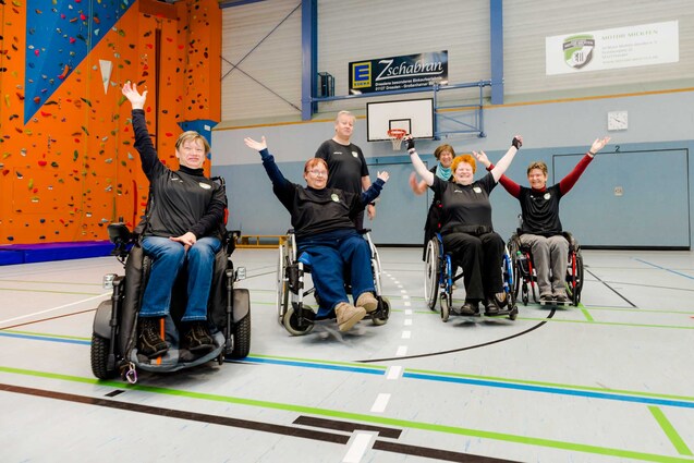 Sechs Personen im Rollstuhl posieren freudig in einer sportlichen Halle mit Kletterwand im Hintergrund.