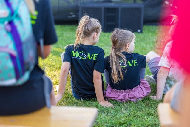 Zwei M&auml;dchen sitzen auf einer Wiese, tragen schwarze T-Shirts mit dem Aufdruck &bdquo;MOVE&ldquo; und blicken auf eine Veranstaltung.