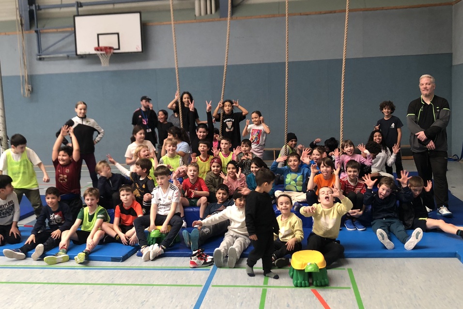 Gruppennahaufnahme von etwa 40 Kindern und Jugendlichen, die auf Sportmatten in einer Turnhalle posieren.