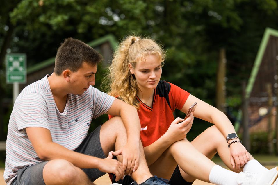 Zwei Jugendliche sitzen im Freien und schauen auf ein Smartphone. Ein Junge in gestreiftem T-Shirt und ein Mädchen in rotem Oberteil.